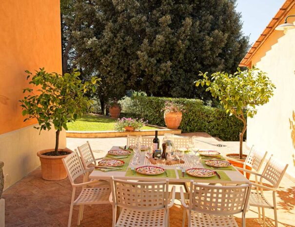 Italian lunch in the garden of Villa Bellavista; white table and chairs with ceramic plates; garden with pine and olive trees in a sunny day - villa rentals by Timeless Tuscany tour operator