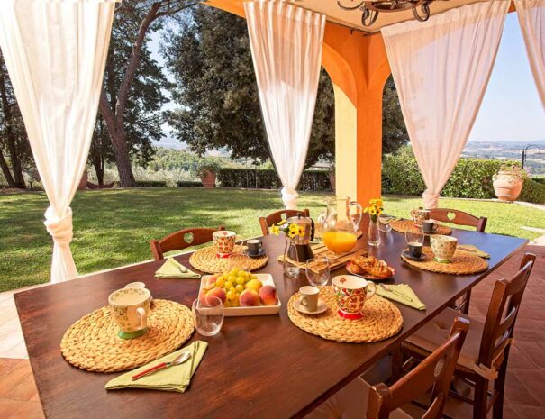 Italian breakfast at Villa Bellavista; table with moka coffee, fruits, bread, marmalade; under porch with white linen shades; garden with pine and olive trees in a sunny day - villa rentals by Timeless Tuscany tour operator