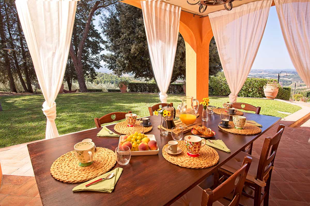 Italian breakfast at Villa Bellavista; table with moka coffee, fruits, bread, marmalade; under porch with white linen shades; garden with pine and olive trees in a sunny day - villa rentals by Timeless Tuscany tour operator